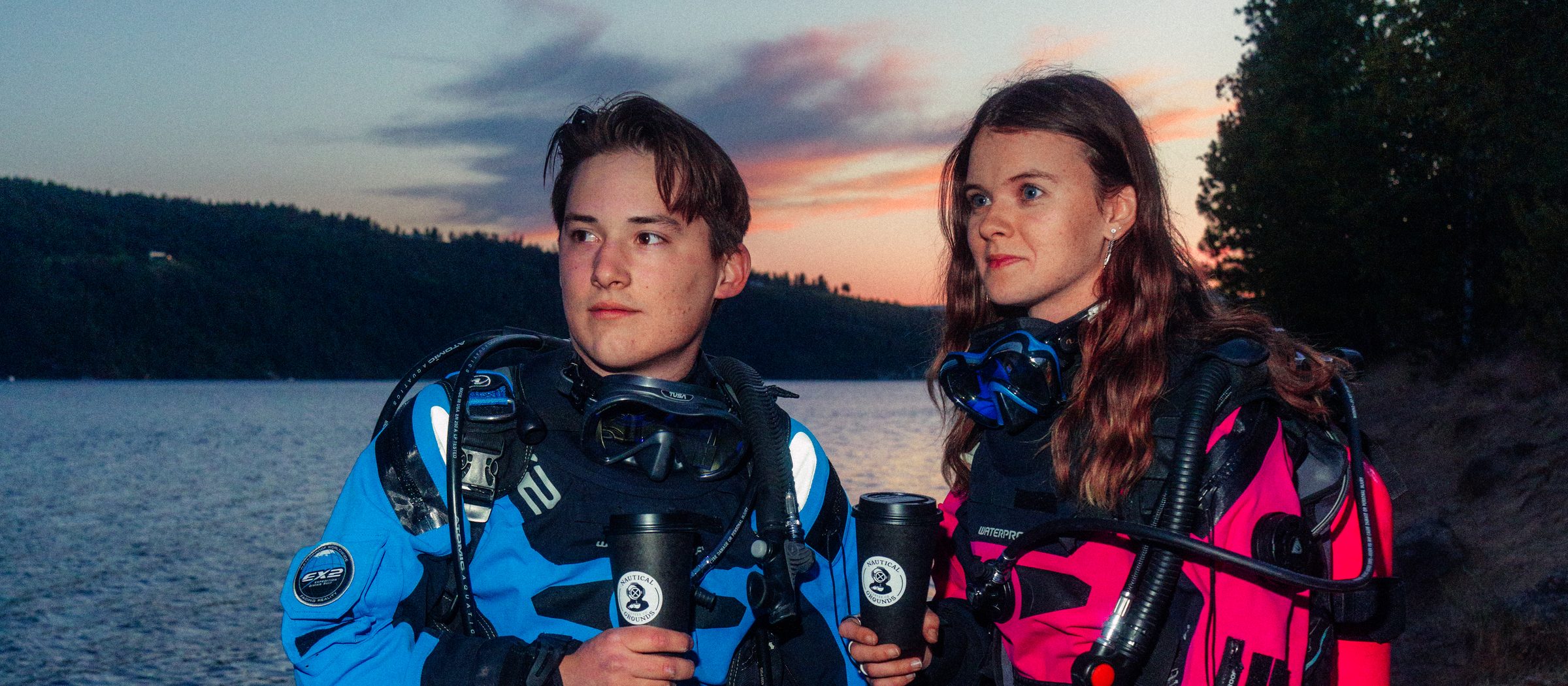 Ethan and Lindsey in dive gear on the shore of Lake Coeur d'Alene at sunset, each holding a Nautical Grounds cup